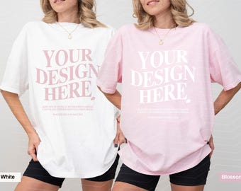 May include: Two short-sleeved crew neck t-shirts, one white and one light pink, each featuring the text "YOUR DESIGN HERE" in a large, bold, serif font, with smaller instructional text below, worn with black shorts and a simple gold necklace.