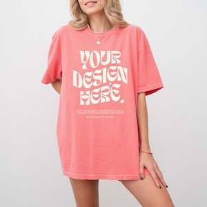 May include: A coral-colored short-sleeve t-shirt featuring the white text "YOUR DESIGN HERE" in a bold, wavy font. Below the main text, smaller white text reads "ADD YOUR DESIGN WITH PHOTOSHOP CANVA OR OTHER EDITING PROGRAM WATERMELON MOCKS."