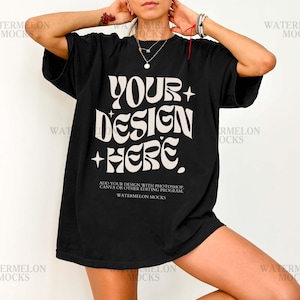 以下が含まれることがあります： レトロな雰囲気の波打つフォントでベージュの「YOUR DESIGN HERE」という文字と、小さな四芒星が描かれた黒の半袖Tシャツ。メインのテキストの下には、編集手順を説明する小さな白いテキストが記載されています。
