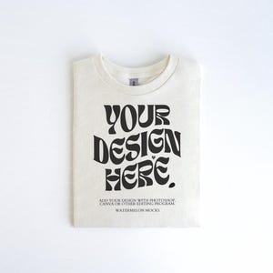 Pu&ograve; includere: Una maglietta bianca con una grafica nera che dice "YOUR DESIGN HERE". Il testo &egrave; in uno stile di font retr&ograve;. La maglietta &egrave; piegata e appoggiata su una superficie bianca. Il testo sotto la grafica dice "ADD YOUR DESIGN WITH PHOTOSHOP, CANVA OR OTHER EDITING PROGRAM. WATERMELON MOCKS."
