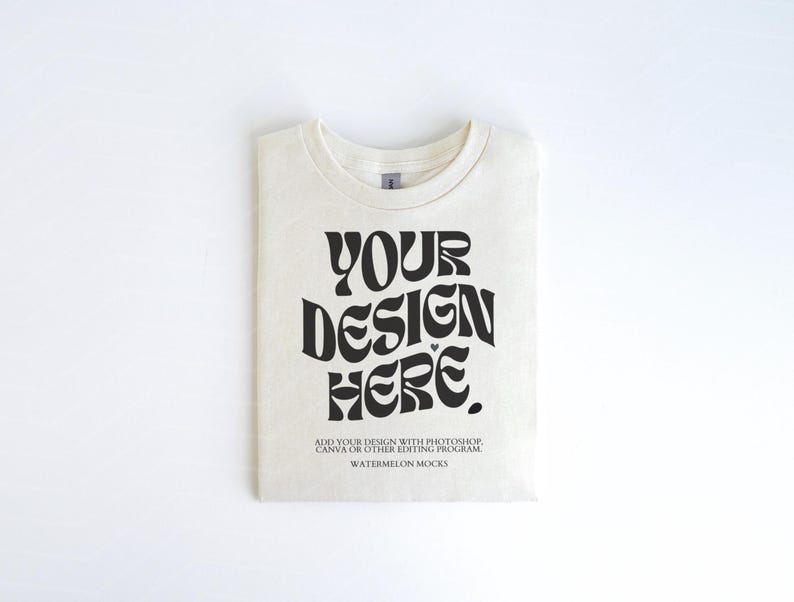 Pu&ograve; includere: Una maglietta bianca con una grafica nera che dice "YOUR DESIGN HERE". Il testo &egrave; in uno stile di font retr&ograve;. La maglietta &egrave; piegata e appoggiata su una superficie bianca. Il testo sotto la grafica dice "ADD YOUR DESIGN WITH PHOTOSHOP, CANVA OR OTHER EDITING PROGRAM. WATERMELON MOCKS."