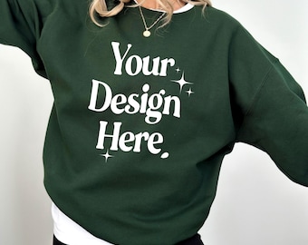 May include: A dark forest green crewneck sweatshirt featuring the white text Your Design Here in a bold serif font, accented by small four-pointed star graphics above and below the text, layered over a white shirt and black shorts.