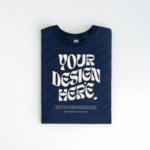 Pu&ograve; includere: Una maglietta blu navy piegata con una grafica bianca che dice "YOUR DESIGN HERE". Il testo &egrave; in uno stile di font retr&ograve;. La maglietta &egrave; su uno sfondo bianco.