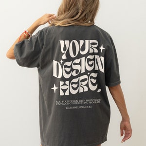 以下が含まれることがあります： 白地に「YOUR+DESIGN+HERE」と書いてある濃いグレーのTシャツ。レトロなフォントスタイルのテキスト。メインテキストの下には「ADD YOUR DESIGN WITH PHOTOSHOP, CANVA OR OTHER EDITING PROGRAM. WATERMELON MOCKS」というテキストが白で印刷されています。