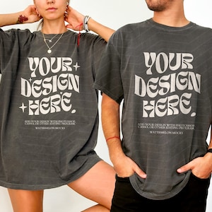 Op de afbeelding: Twee mensen dragen oversized, donkergrijze T-shirts met de tekst "YOUR DESIGN HERE" in wit. De tekst is in een retro, psychedelisch lettertype. De shirts zijn een mockup voor het toevoegen van een eigen ontwerp.