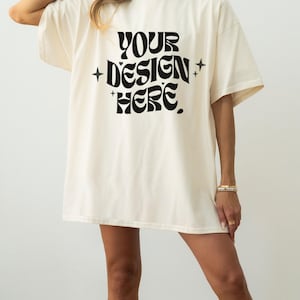 Pode incluir: Uma t-shirt oversize de cor creme com o texto preto "YOUR DESIGN HERE" impresso na parte frontal. O texto apresenta um tipo de letra ondulado e em negrito, ladeado por pequenos ícones de estrelas de quatro pontas em ambos os lados.