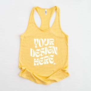 May include: A bright yellow racerback tank top featuring the white text YOUR DESIGN HERE printed in a wavy, bold font across the center. The garment has a scoop neckline and a sleeveless design with a subtle, repeating chevron pattern.