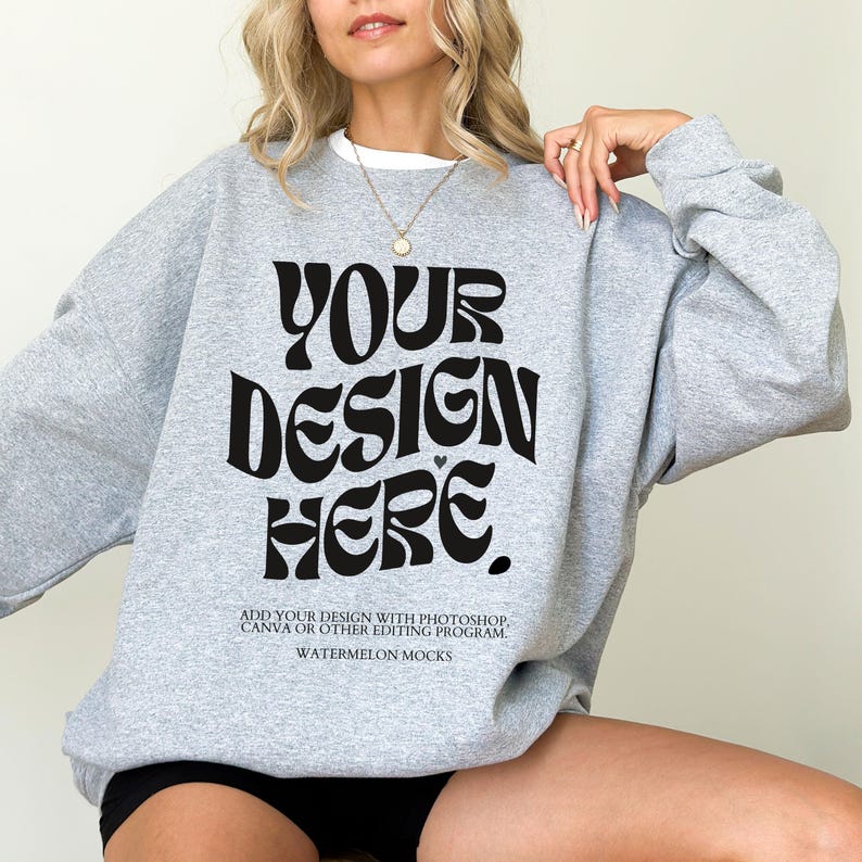May include: A grey sweatshirt with the text "YOUR DESIGN HERE" in black. The text is in a retro font style. The text below the design reads "ADD YOUR DESIGN WITH PHOTOSHOP, CANVA OR OTHER EDITING PROGRAM. WATERMELON MOCKS."