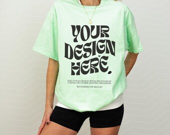 Puede incluir: Una camiseta de manga corta de color verde claro con el texto en negro YOUR DESIGN HERE, seguido de un texto más pequeño que dice ADD YOUR DESIGN WITH PHOTOSHOP, CANVA OR OTHER EDITING PROGRAM y WATERMELON MOCKS abajo.