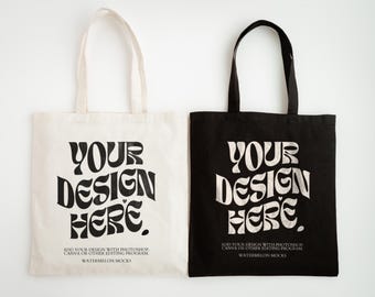 May include: Two rectangular tote bags, one off-white and one black, each featuring two shoulder straps. The text "YOUR DESIGN HERE" is printed in a large, wavy black font, with smaller text below reading "ADD YOUR DESIGN WITH PHOTOSHOP, CANVA OR OTHER EDITING PROGRAM" and "WATERMELON MOCKS."