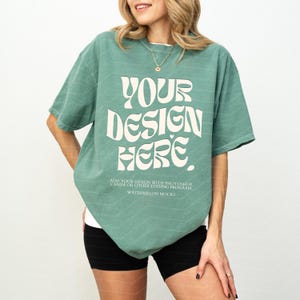 Pode incluir: Uma t-shirt de manga curta verde-sálvia com o texto branco "YOUR DESIGN HERE." e um texto menor abaixo que diz "ADD YOUR DESIGN WITH PHOTOSHOP, CANVA OR OTHER EDITING PROGRAM." e "WATERMELON MOCKS." A t-shirt tem um corte largo e descontraído.