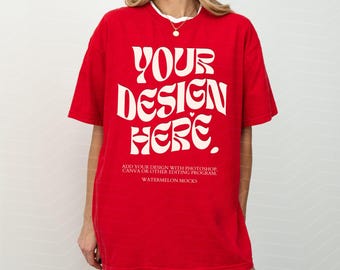 May include: A bright red short-sleeved t-shirt featuring large, bold, cream-colored text that reads YOUR DESIGN HERE. Below this, smaller text states ADD YOUR DESIGN WITH PHOTOSHOP, CANVA OR OTHER EDITING PROGRAM. WATERMELON MOCKS.