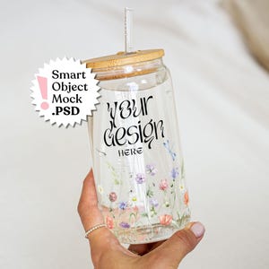 Puede incluir: Un vaso de cristal transparente con tapa de bambú y pajita de cristal. El vaso presenta un estampado floral con pequeñas flores de colores y el texto negro "YOUR DESIGN HERE". Incluye un gráfico de estrella blanco con "Smart Object .PSD".