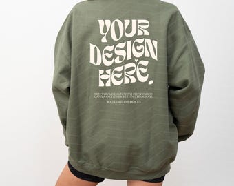 Op de afbeelding: Een olijfgroene sweater met ronde hals en de witte tekst "YOUR DESIGN HERE" in een groot, golvend lettertype op de achterkant. Onder de hoofdtekst staat in kleinere witte letters "ADD YOUR DESIGN WITH PHOTOSHOP, CANVA OR OTHER EDITING PROGRAM" en "WATERMELON MOCKS."