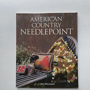 May include: A book titled American Country Needlepoint by Jim Williams. The cover features a brown wooden background, a needlepoint pillow with a rooster, a floral patterned throw blanket draped over a chair, and two small baskets of berries.