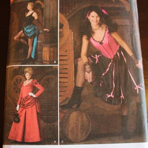 May include: Simplicity sewing pattern featuring three costume designs. The costumes include a black and teal saloon girl outfit, a pink and black dress with bows, and a red Victorian gown. The pattern includes sizes 14-20.