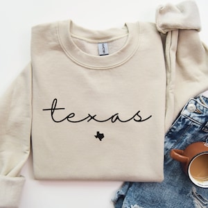 May include: A beige sweatshirt with the word "texas" written in black cursive script with a small outline of the state of Texas underneath.