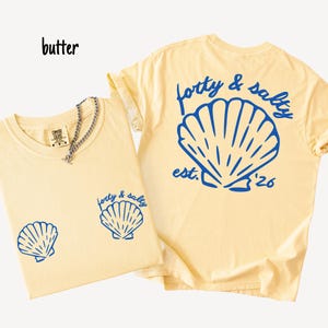 May include: Two butter-colored t-shirts with blue seashell graphics. One shirt has a small seashell graphic on the front and the text "forty & salty" with a seashell graphic on the back. The other shirt has two small seashell graphics on the front.