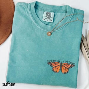 May include: Seafoam green t-shirt with a butterfly graphic and the text "busy growing a tiny human." The shirt has a crew neck and short sleeves. A gold necklace with a small pendant is draped across the shirt.