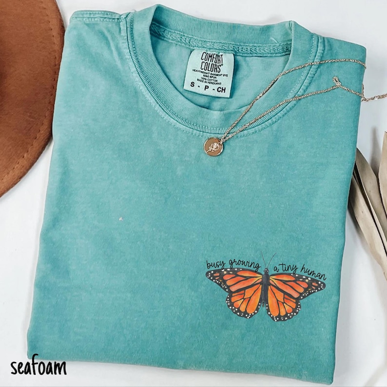 May include: Seafoam green t-shirt with a butterfly graphic and the text "busy growing a tiny human." The shirt has a crew neck and short sleeves. A gold necklace with a small pendant is draped across the shirt.
