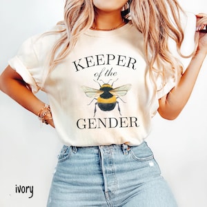 May include: A white t-shirt with the text "KEEPER of the GENDER" and a cartoon bumblebee illustration. The shirt is on a model wearing blue jeans.