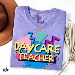 May include: A violet-colored t-shirt with the words "DAYCARE TEACHER" in a colorful 80s-style design. The shirt has a crew neck and short sleeves. A gold necklace with a pendant is draped across the shirt.