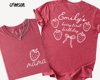 May include: Two crimson-colored t-shirts. One shirt has a white strawberry graphic and the word "mama". The other shirt has the text "Emily's berry first birthday" with strawberry graphics. A silver chain necklace is on the first shirt.