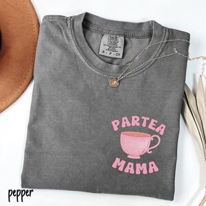 Puede incluir: Una camiseta gris Comfort Colors con las palabras "Partea Mama" y un gr&aacute;fico de taza de t&eacute; rosa. Un collar plateado con un colgante est&aacute; drapeado sobre la camisa. La palabra "pepper" est&aacute; impresa en la esquina inferior izquierda.