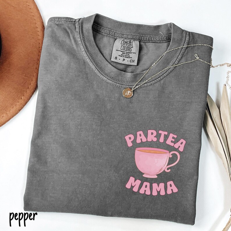 Puede incluir: Una camiseta gris Comfort Colors con las palabras "Partea Mama" y un gr&aacute;fico de taza de t&eacute; rosa. Un collar plateado con un colgante est&aacute; drapeado sobre la camisa. La palabra "pepper" est&aacute; impresa en la esquina inferior izquierda.