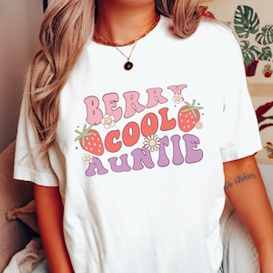 May include: White t-shirt with a pink, red, and purple graphic that says "Berry Cool Auntie" with strawberries and flowers.