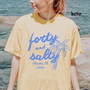 May include: Butter-colored t-shirt with the text "forty and salty" in blue, along with a palm tree graphic. Below the text is "Miami, FL 2026". The shirt is a casual style.