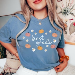 May include: A light blue t-shirt with the text "first grade" in white surrounded by a circle of small white daisies and orange and pink pumpkins.
