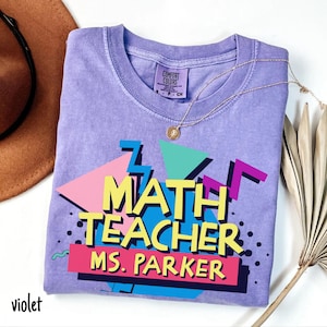May include: A violet Comfort Colors t-shirt with a retro design that reads "MATH TEACHER MS. PARKER" in colorful block letters. A gold necklace with a pendant rests on the shirt. The word "violet" is printed in the bottom left corner.