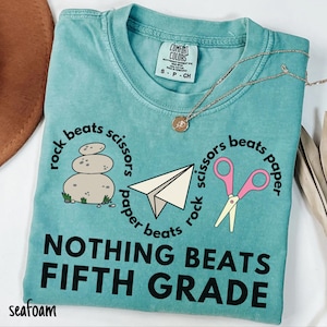 May include: Seafoam green t-shirt with the text "Rock beats scissors, scissors beats paper, paper beats rock. Nothing beats fifth grade." The shirt features illustrations of a stack of rocks, a paper airplane, and pink scissors.