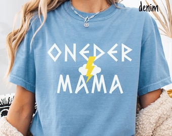 Op de afbeelding: Lichtblauw T-shirt met de witte woorden "ONE DER MAMA", versierd met een gele bliksemschicht en een wolk. De woorden "washed denim" staan rechtsboven. Het T-shirt is een casual, comfortabele stijl.