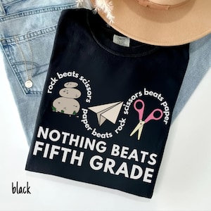 May include: Black t-shirt with a graphic design featuring a paper airplane, scissors, and stacked stones, with the text "Rock beats scissors, scissors beats paper, paper beats rock" and "Nothing beats Fifth Grade."