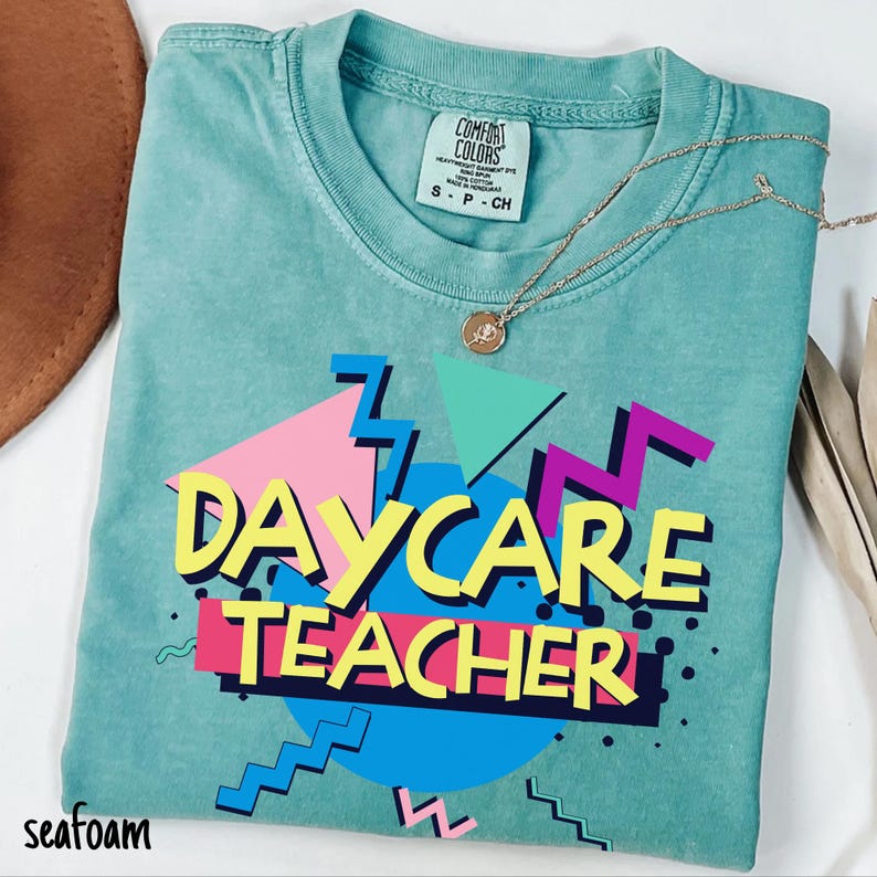 May include: Seafoam green t-shirt with the words "DAYCARE TEACHER" in a retro design. The design includes pink, yellow, blue, and purple geometric shapes. A gold necklace with a pendant rests on the shirt. The shirt is labeled "Comfort Colors".