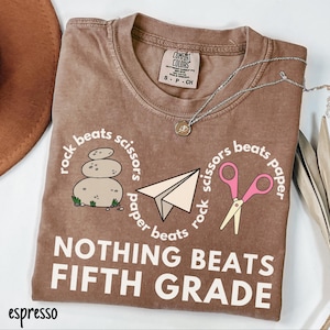May include: Espresso-colored t-shirt with the text "NOTHING BEATS FIFTH GRADE." The shirt features a rock, paper, scissors design with a stack of stones, a paper airplane, and pink scissors. A silver necklace with a pendant is on the shirt.