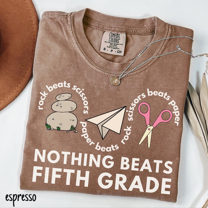 May include: Espresso-colored t-shirt with the text "NOTHING BEATS FIFTH GRADE." The shirt features a rock, paper, scissors design with a stack of stones, a paper airplane, and pink scissors. A silver necklace with a pendant is on the shirt.