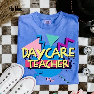 May include: A blue t-shirt with the words "DAYCARE TEACHER" in a colorful 80s-style design. The shirt is paired with white sneakers. The shirt is made of a soft material and is perfect for a daycare teacher.