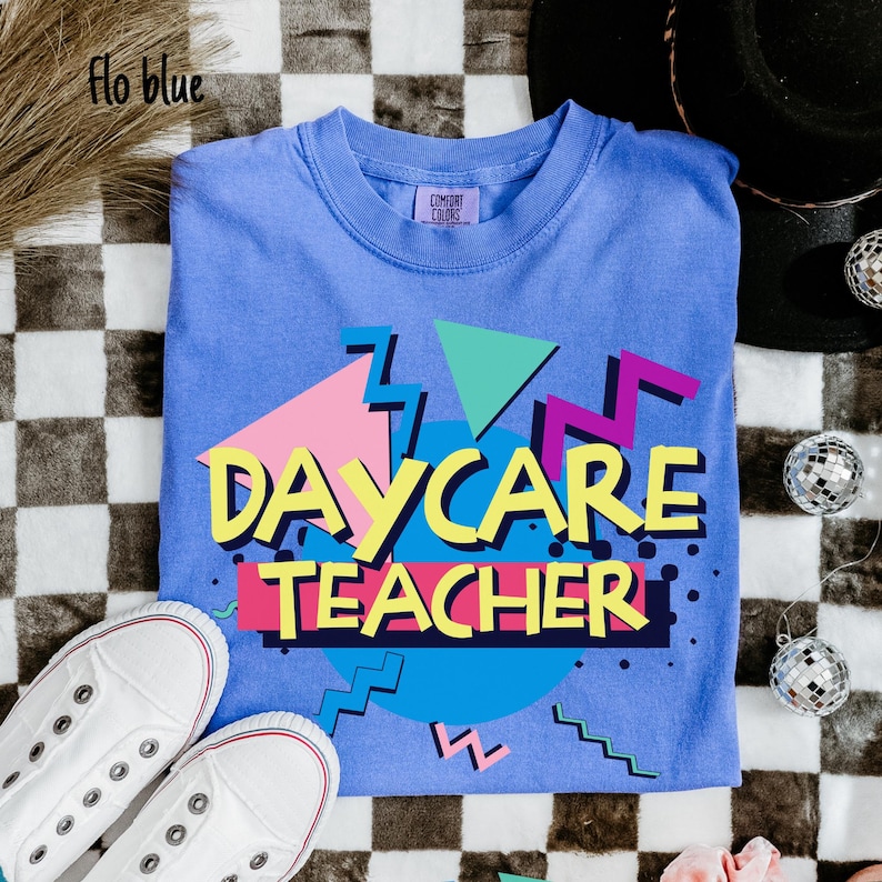 May include: A blue t-shirt with the words "DAYCARE TEACHER" in a colorful 80s-style design. The shirt is paired with white sneakers. The shirt is made of a soft material and is perfect for a daycare teacher.