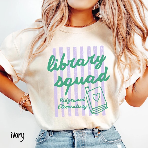 以下が含まれることがあります： アイボリー色のTシャツ。緑色の筆記体で「library squad」と書かれ、ストライプの背景が特徴。その下に、ハートの描かれた本の緑色の輪郭と「Ridgewood Elementary」の文字が配置されています。