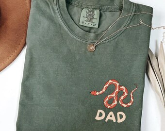 May include: Moss green t-shirt with a red and white snake graphic and the word "DAD". The shirt has a crew neck and short sleeves. A silver necklace with a pendant is on the shirt.