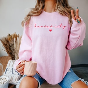 May include: A pink sweatshirt with the text "kansas city" in red script with a small red heart above the text.