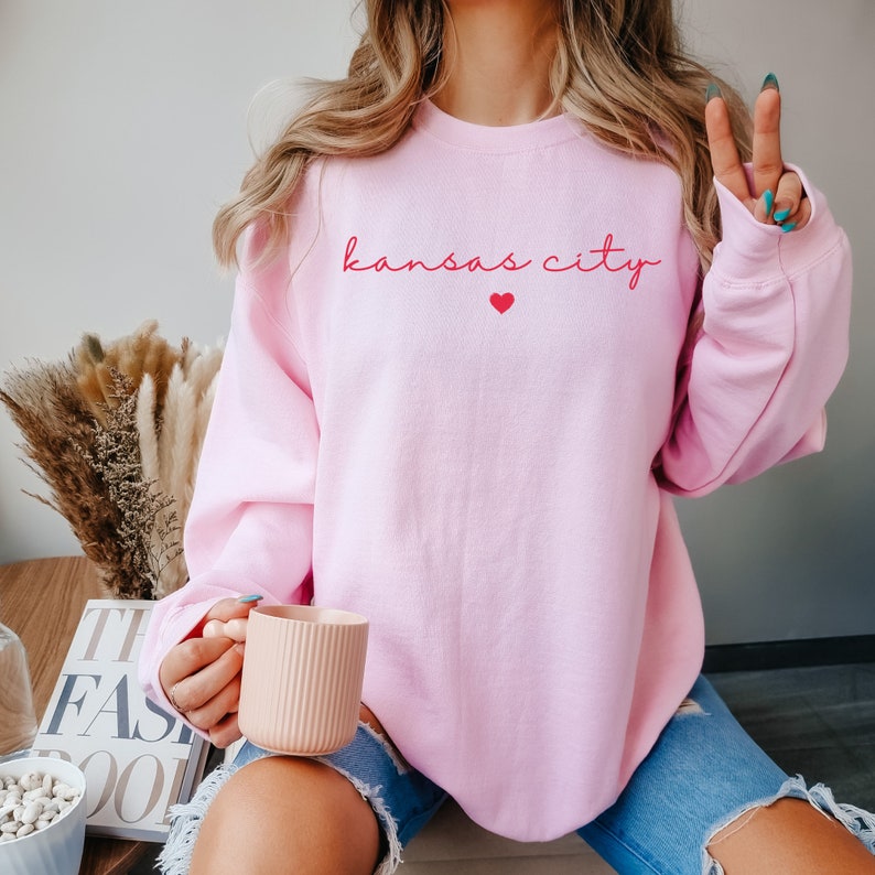 May include: A pink sweatshirt with the text "kansas city" in red script with a small red heart above the text.