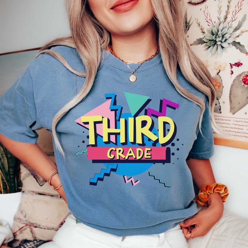 Op de afbeelding: Blauw T-shirt met een kleurrijke "THIRD GRADE" afbeelding. Het ontwerp bevat geometrische vormen in roze, geel, turquoise en blauw, omlijnd met zwart. Het shirt heeft korte mouwen en een ronde hals.
