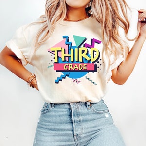 Op de afbeelding: Cr&egrave;mekleurig T-shirt met een retro ontwerp met de woorden "THIRD GRADE" in geel, omlijnd met zwart. Het ontwerp bevat geometrische vormen in roze, teal en paars, met een blauwe cirkel en een roze en zwarte rechthoek.
