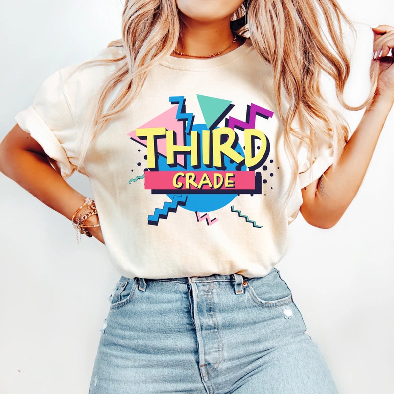 Op de afbeelding: Cr&egrave;mekleurig T-shirt met een retro ontwerp met de woorden "THIRD GRADE" in geel, omlijnd met zwart. Het ontwerp bevat geometrische vormen in roze, teal en paars, met een blauwe cirkel en een roze en zwarte rechthoek.