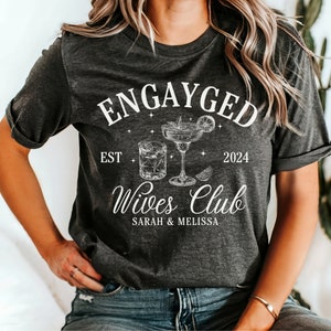 May include: A gray t-shirt with the text "Engayged Est 2024 Wives Club Sarah & Melissa" and a drawing of two cocktails.