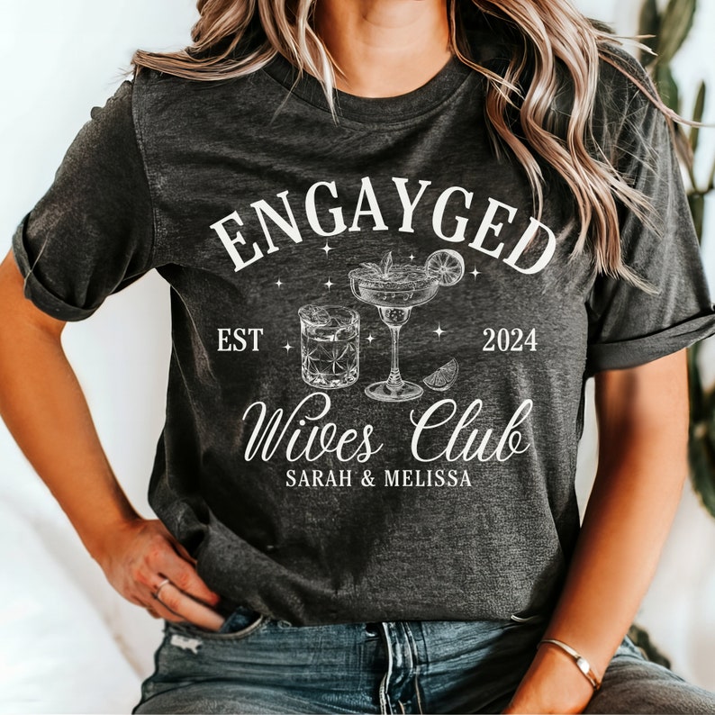 May include: A gray t-shirt with the text "Engayged Est 2024 Wives Club Sarah & Melissa" and a drawing of two cocktails.